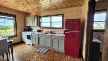 A compact kitchen with wooden ceiling, a red refrigerator, and a window overlooking a rural landscape.