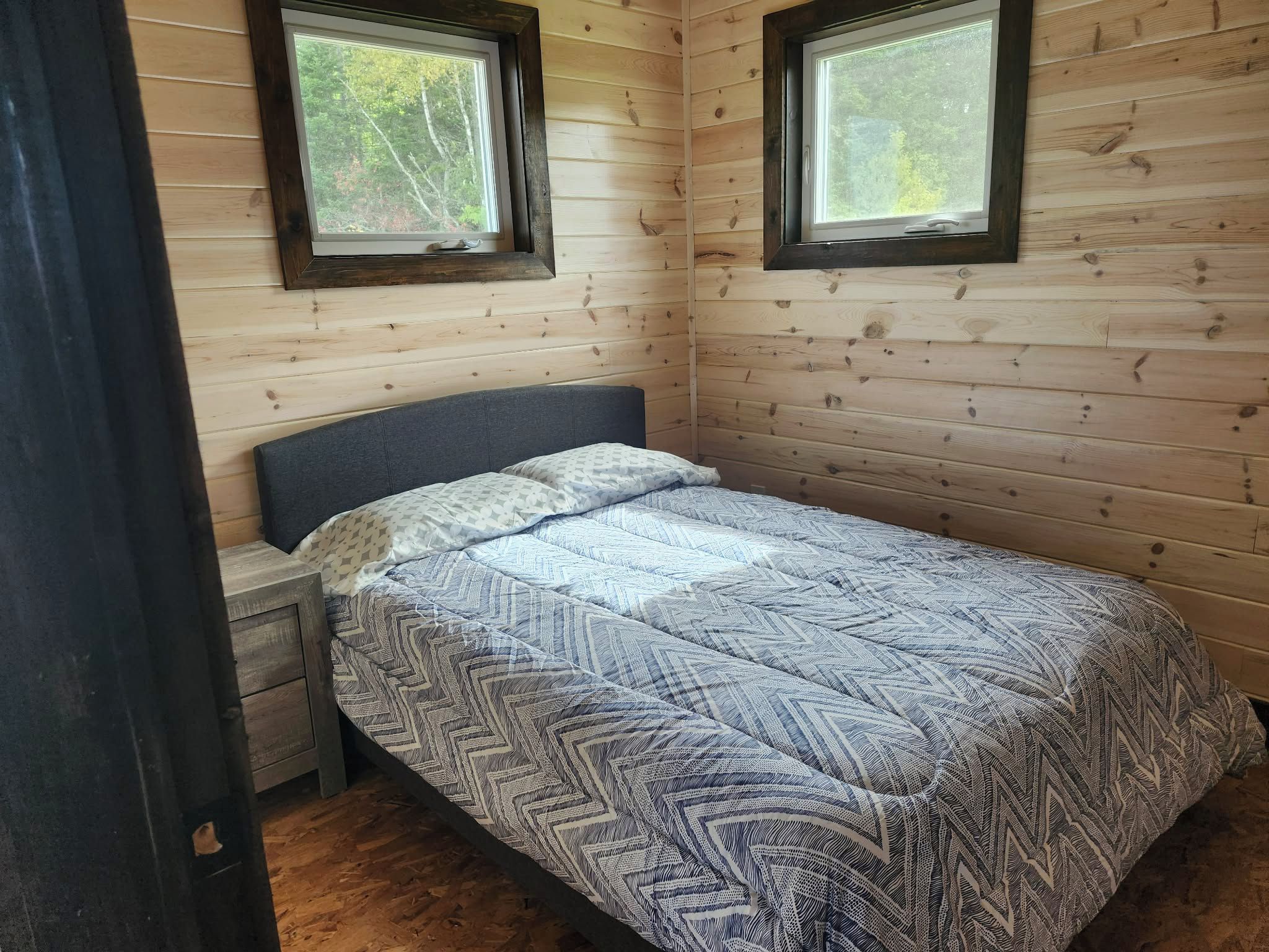 A single bed with a patterned bedspread in a room with wooden walls and two windows.