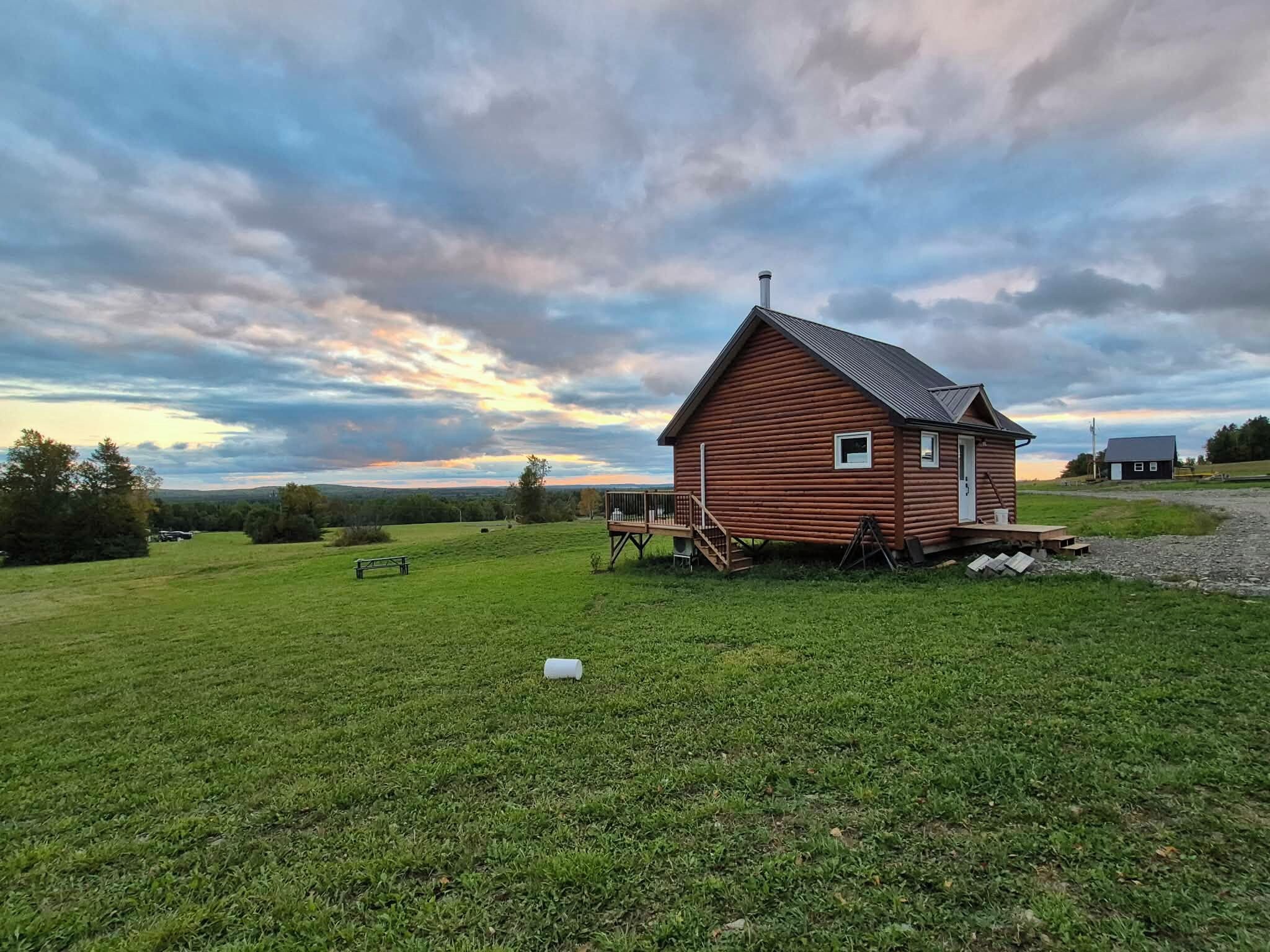A small wooden cabin with a porch, surrounded by a grassy field and a cloudy sky.