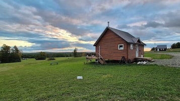 A small wooden cabin with a porch, surrounded by a grassy field and a cloudy sky.