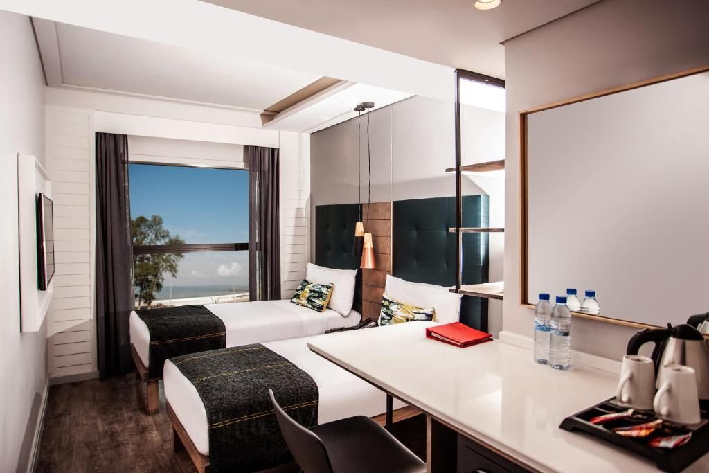 Een hotelkamer met twee bedden, een bureau, een stoel, een groot raam en uitzicht op zee.