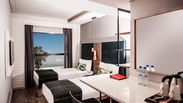 Een hotelkamer met twee bedden, een bureau, een stoel, een groot raam en uitzicht op zee.
