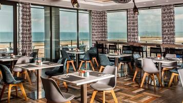 Een restaurant met tafels en stoelen klaar voor diners, grote ramen met uitzicht op zee, en gordijnen met een patroon.