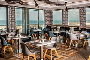 Een restaurant met tafels en stoelen klaar voor diners, grote ramen met uitzicht op zee, en gordijnen met een patroon.