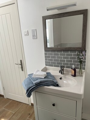 Une salle de bain avec un lavabo blanc, un miroir avec un cadre en bois, un porte-serviettes avec des serviettes pliées et un flacon de savon liquide.