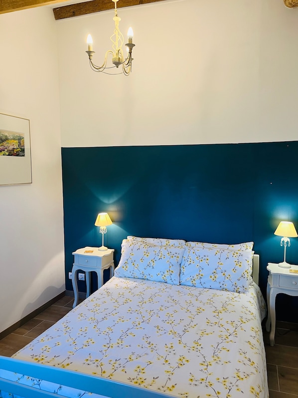 A bedroom with a bed, two bedside tables with lamps, a chandelier, and a wall-mounted picture.