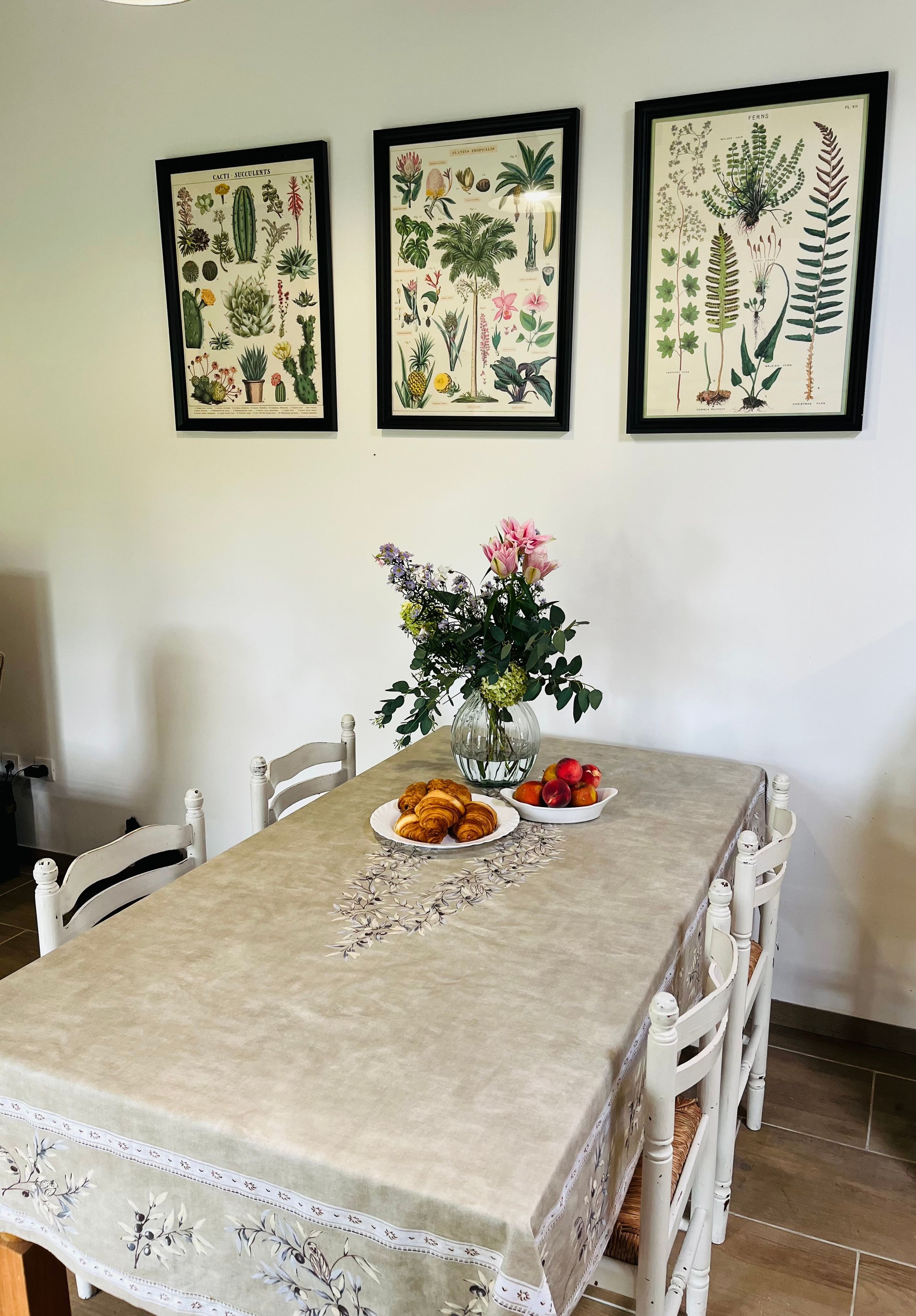 Una taula de menjador amb un mantell floral, rodejada de cadires blanques, amb un ram de flors i plats de menjar.