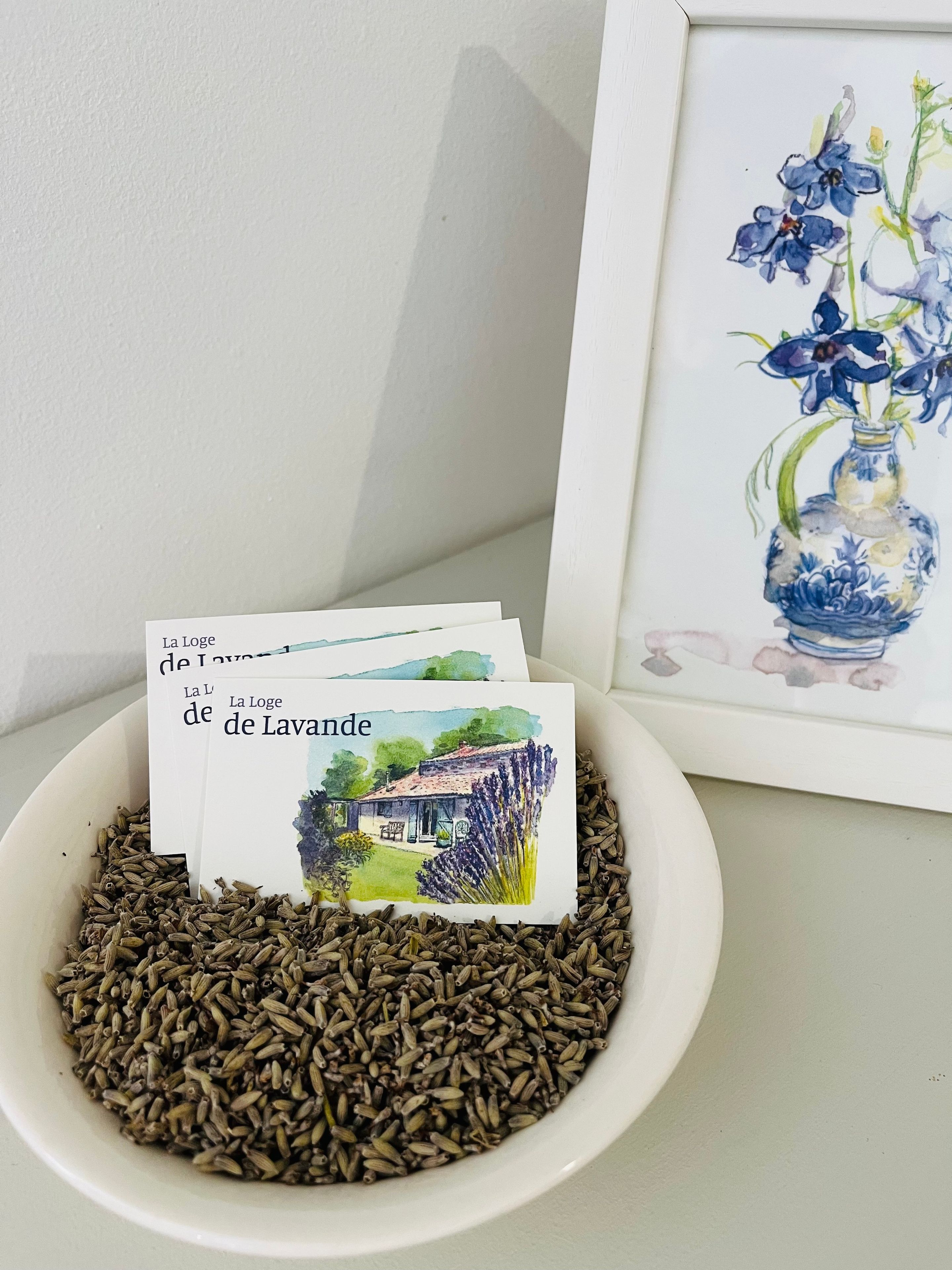 Un tazón de semillas de comino con un cuadro acuarela enmarcado de flores de lavanda y una postal de una casa con el texto 'La Loge de Lavande'.
