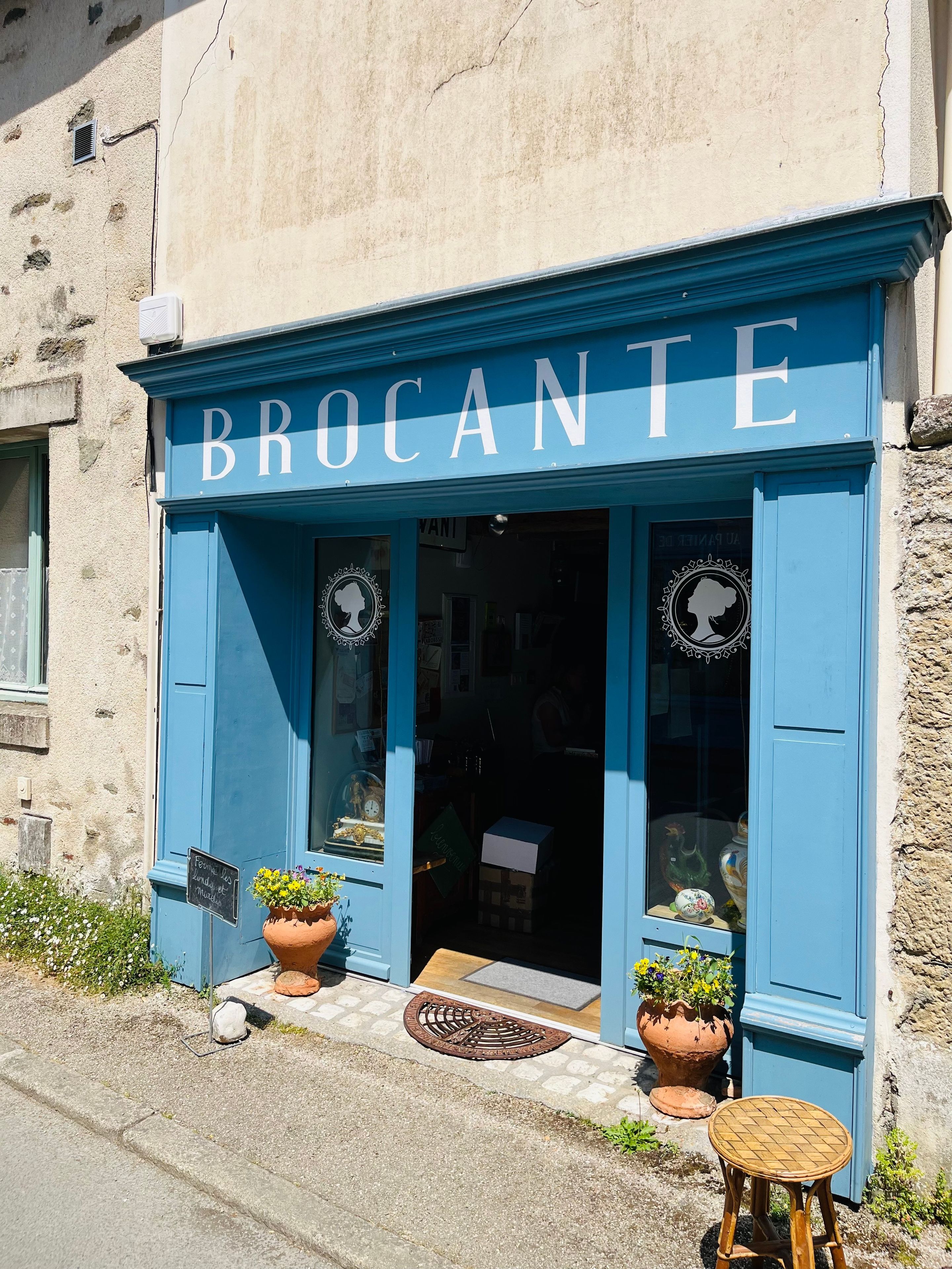 Ein Ladenlokal mit dem Schild 'BROCANTE' und zwei Blumentöpfen vor der Tür.