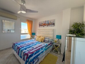 Ein Hotelzimmer mit einem Bett, einem Nachttisch, einem Spiegel, einem Deckenventilator, einem Fenster mit Vorhängen, einem Gemälde und einer Topfpflanze.