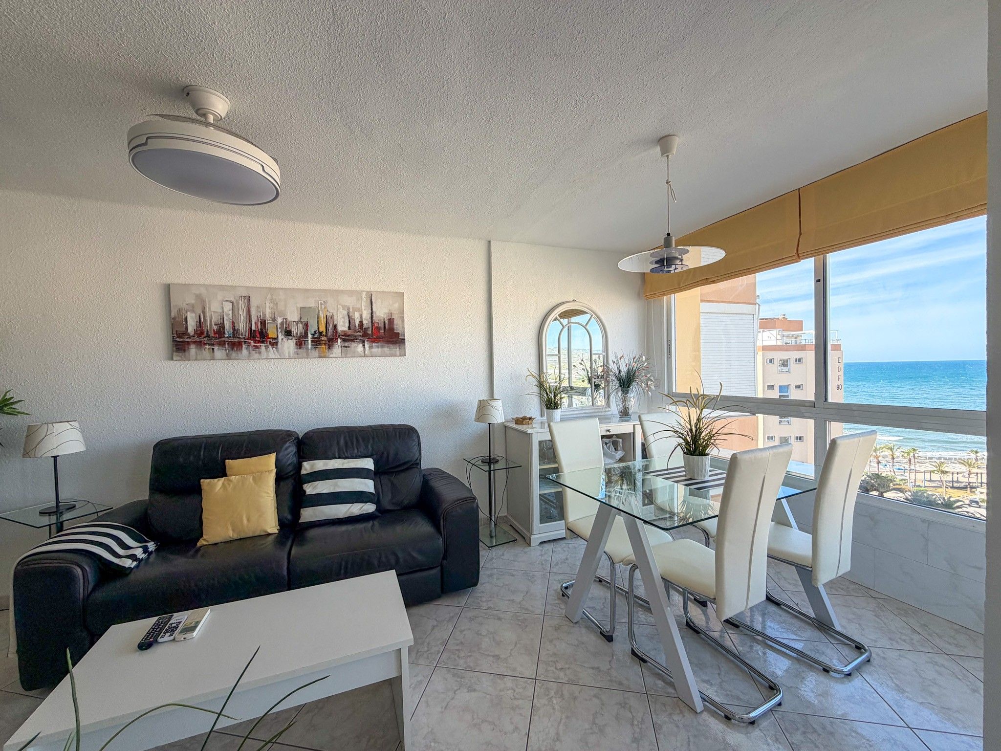 A modern living room with a black leather sofa, a glass dining table, and a cityscape painting on the wall.