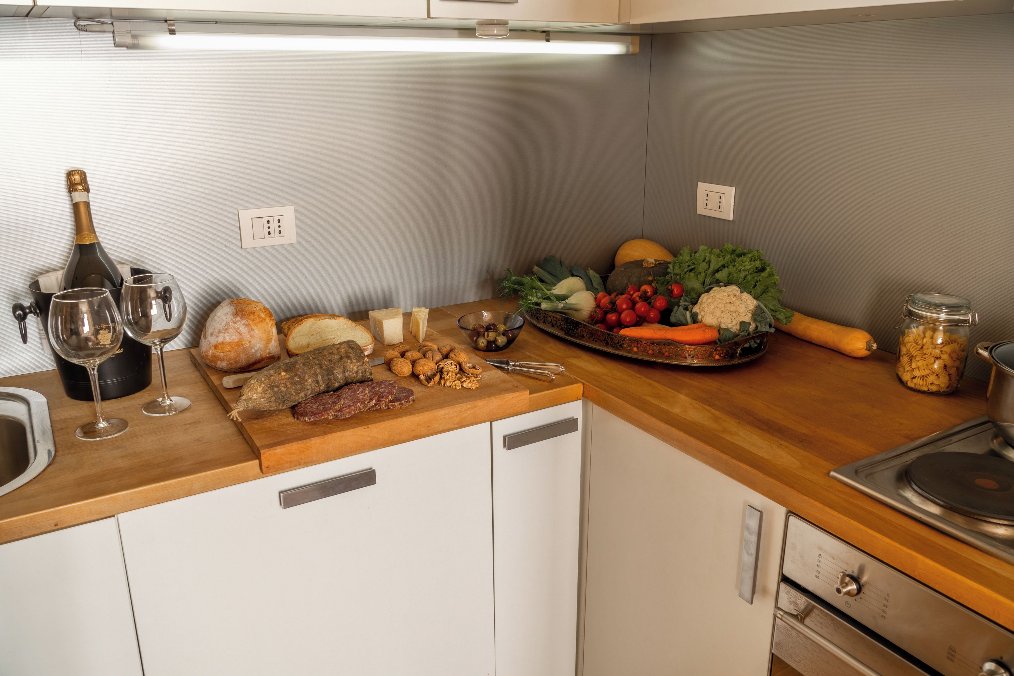 Una cucina con un piano di lavoro in legno, una bottiglia di vino, bicchieri, pane, formaggio, noci e diversi tipi di verdure.
