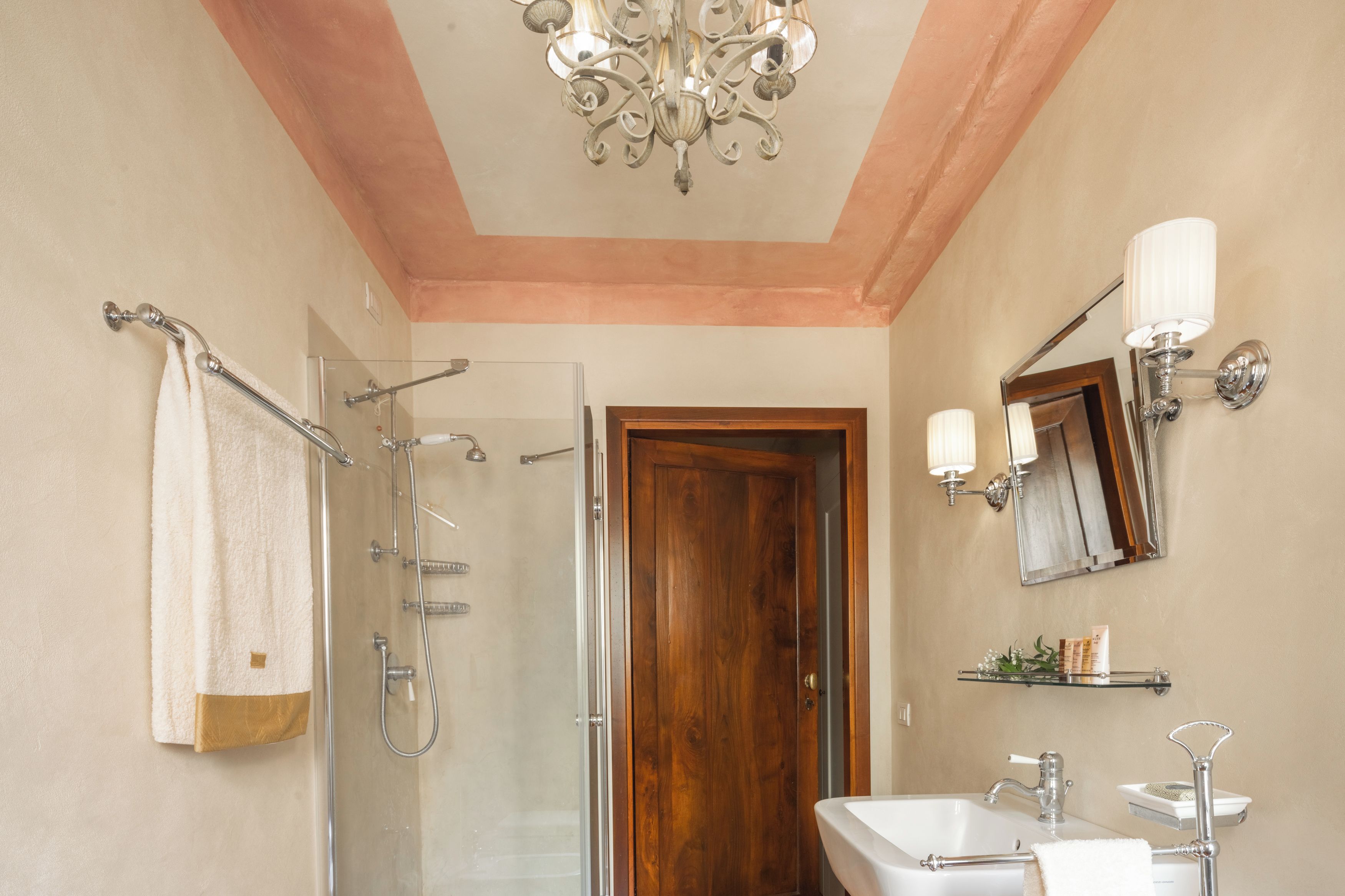 A bathroom with a glass shower enclosure, a wooden door, a white sink, and a wall-mounted mirror with lights.