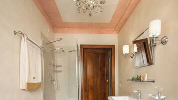 A bathroom with a glass shower enclosure, a wooden door, a white sink, and a wall-mounted mirror with lights.