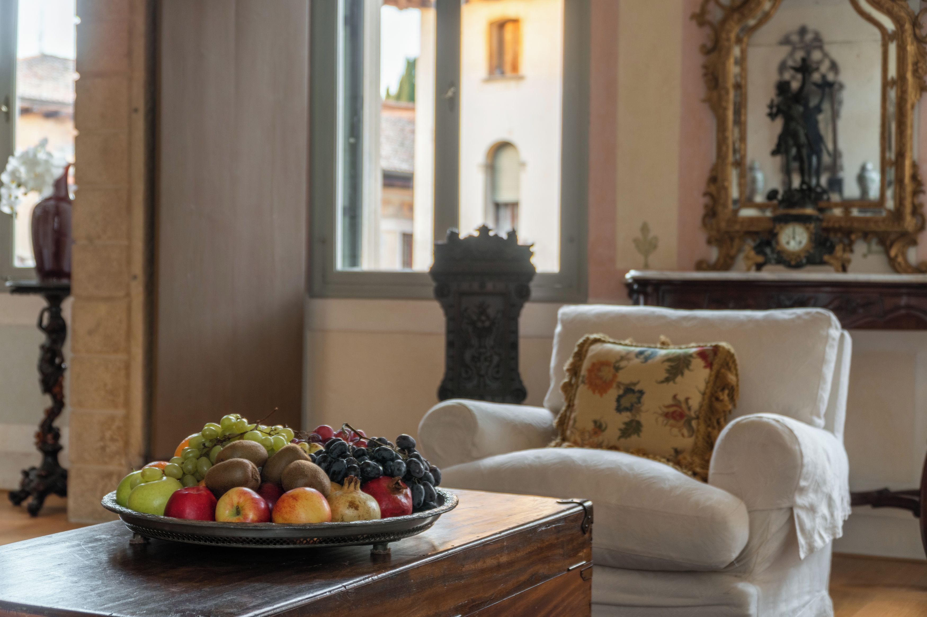 Une pièce avec un fauteuil blanc, une table en bois sur laquelle se trouve un panier de fruits, et une vue sur des immeubles par la fenêtre.