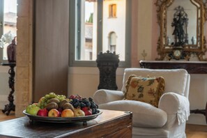 Una habitación con un sillón blanco, una mesa de madera con fruteros, y vistas a edificios a través de una ventana.