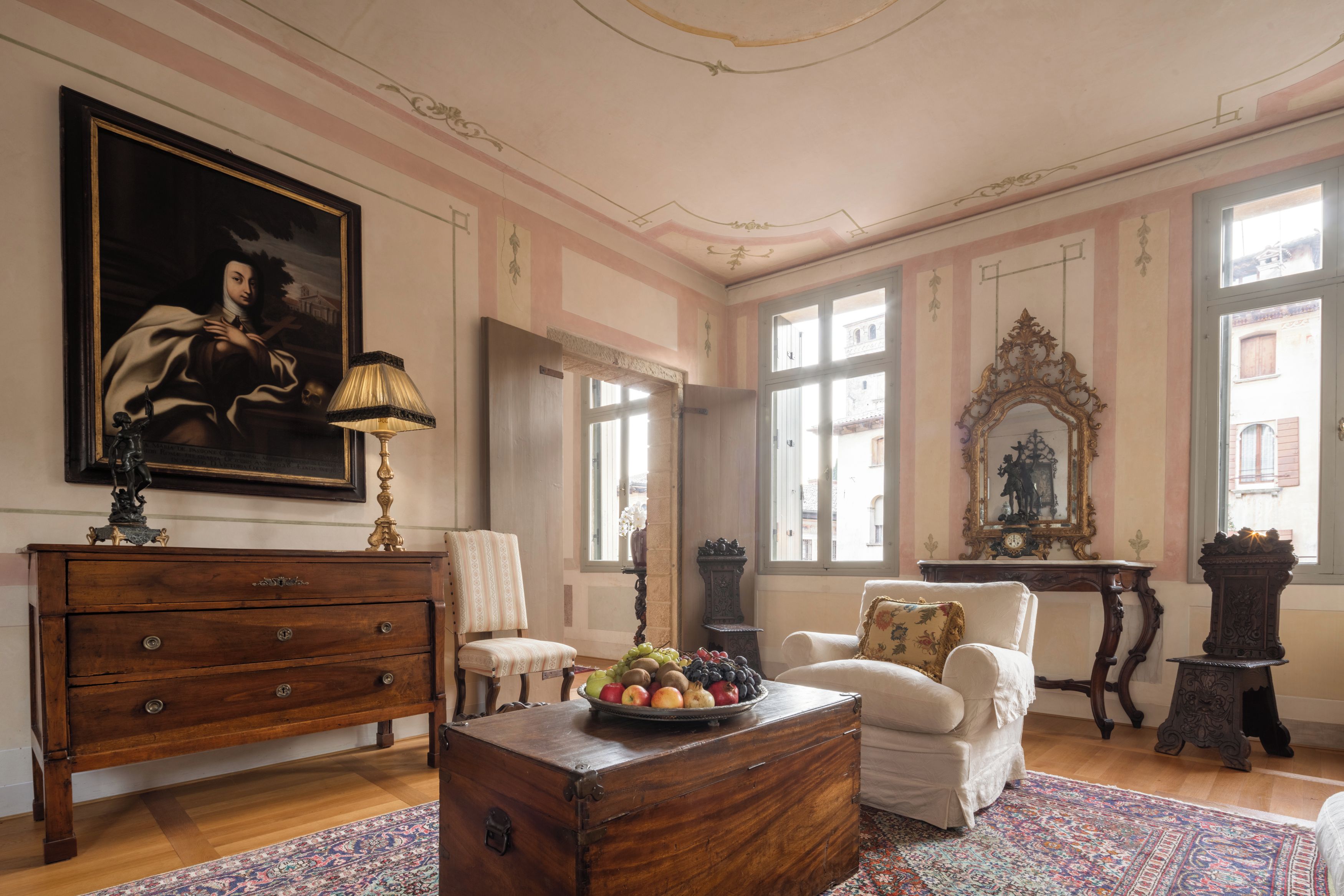Une pièce comprenant un coffre en bois, une chaise, une table avec des fruits, une lampe et un grand tableau accroché au mur.