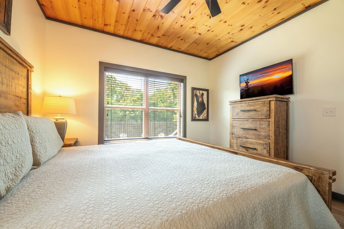 A bedroom with a large bed, a wooden ceiling, a window with blinds, a wooden dresser, and a painting on the wall.