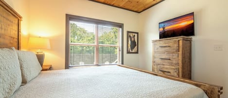 A bedroom with a large bed, a wooden ceiling, a window with blinds, a wooden dresser, and a painting on the wall.