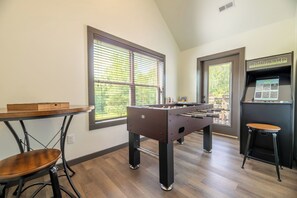 A room with a foosball table, a bar area with a foosball machine, and a window with blinds. - 🌰 Acorn Lodge: Resort Views, Hot Tub, Fire Pit, & Mini Golf, Sleeps 12 (Sevierville)