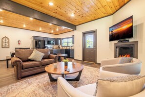 A modern living room with a fireplace, a television, and a kitchen in the background. - 🌰 Acorn Lodge: Resort Views, Hot Tub, Fire Pit, & Mini Golf, Sleeps 12 (Sevierville)