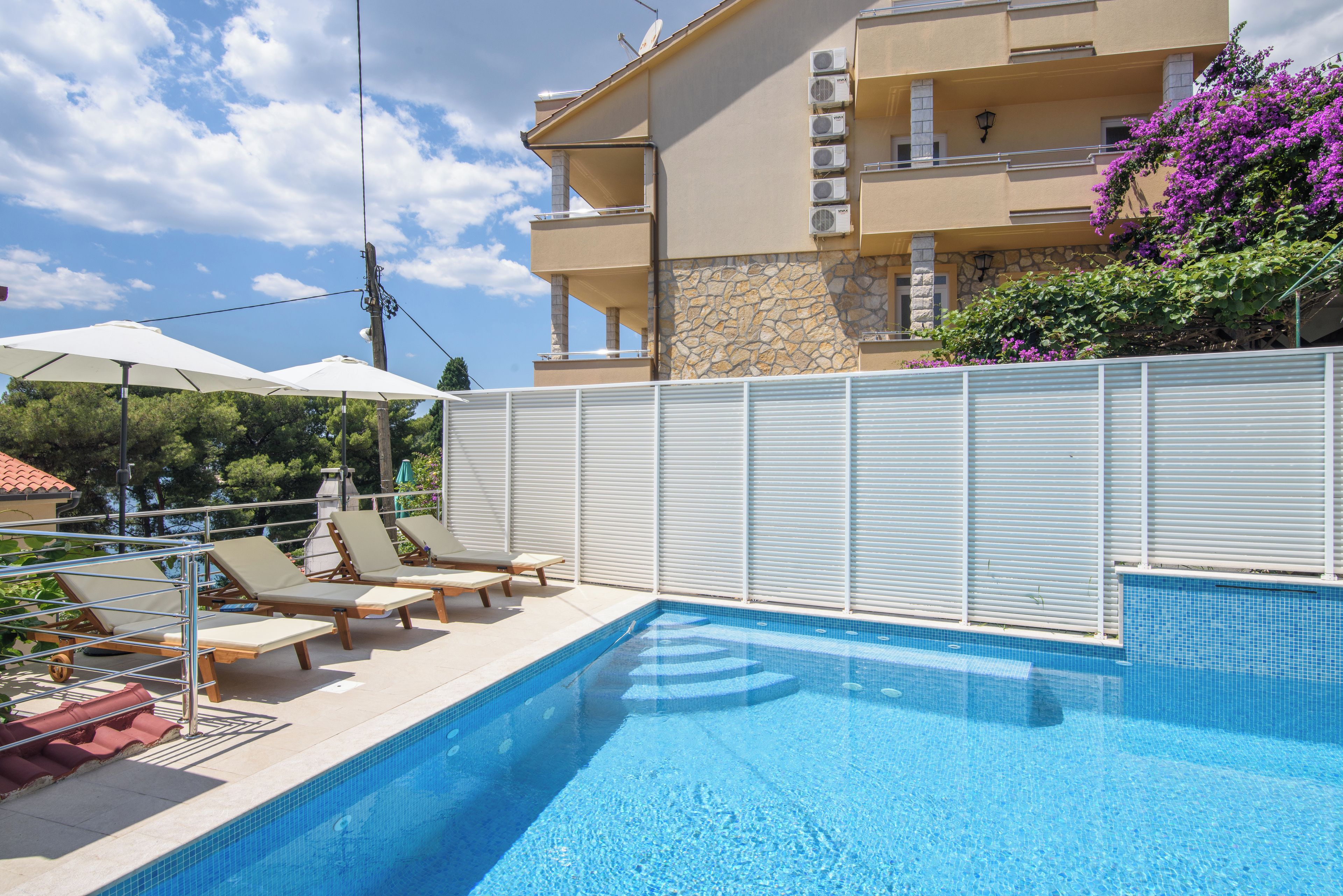 Ein Pool mit Liegestühlen und Sonnenschirmen, umgeben von einem weißen Zaun, mit einem Gebäude im Hintergrund.