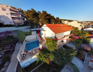 A two-story house with a swimming pool, surrounded by greenery and neighboring buildings. - A3 – 90 m2 apt with shared pool, big terrace and garden, just 50m from the beach (Mastrinka)
