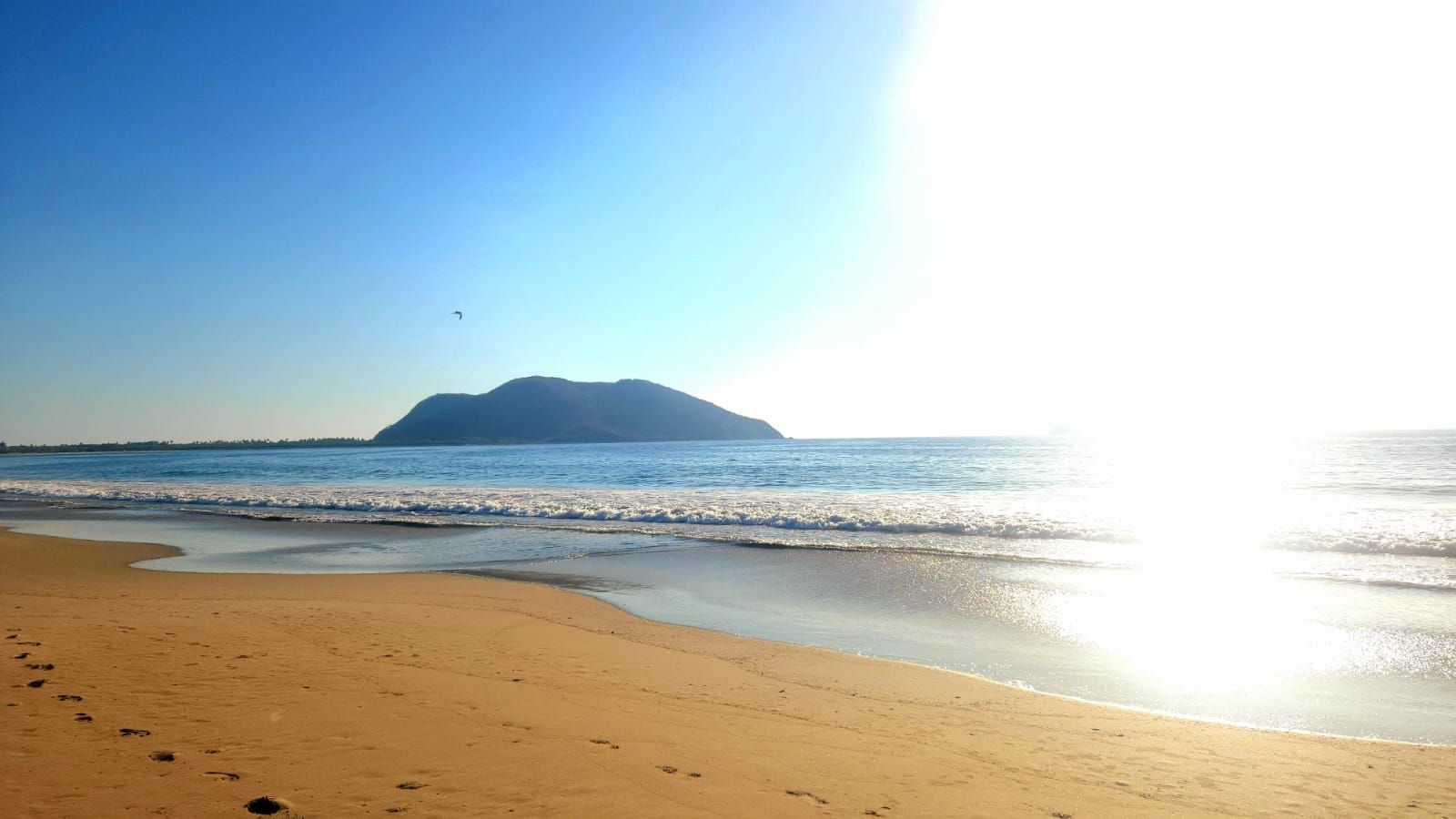 Une plage avec un ciel dégagé, une mer calme et une montagne qui se profile à l’horizon.
