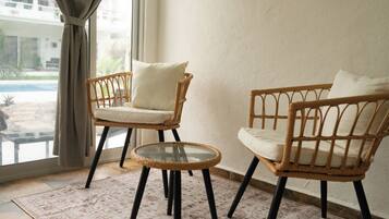Deux fauteuils en osier avec des coussins, une table en verre et un tapis à motifs dans une pièce dotée d’une grande fenêtre.