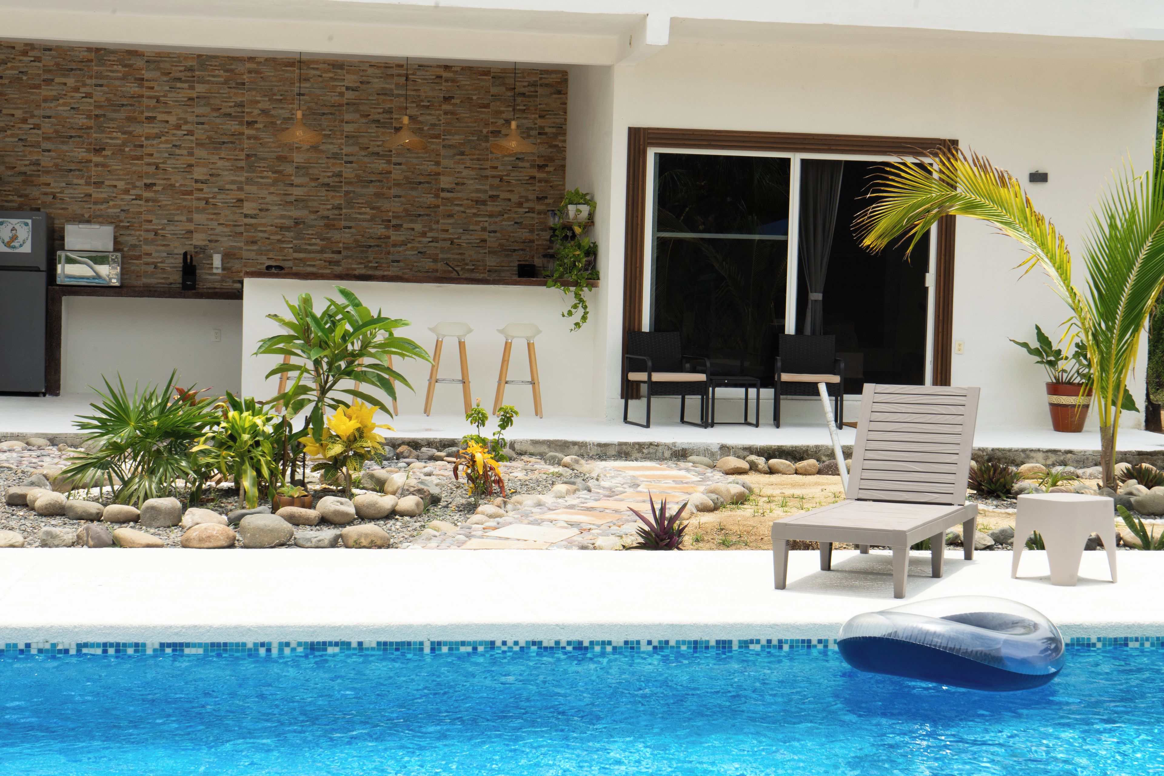 A pool area with lounge chairs, a small garden with rocks and plants, and a modern building with a stone wall and large windows.