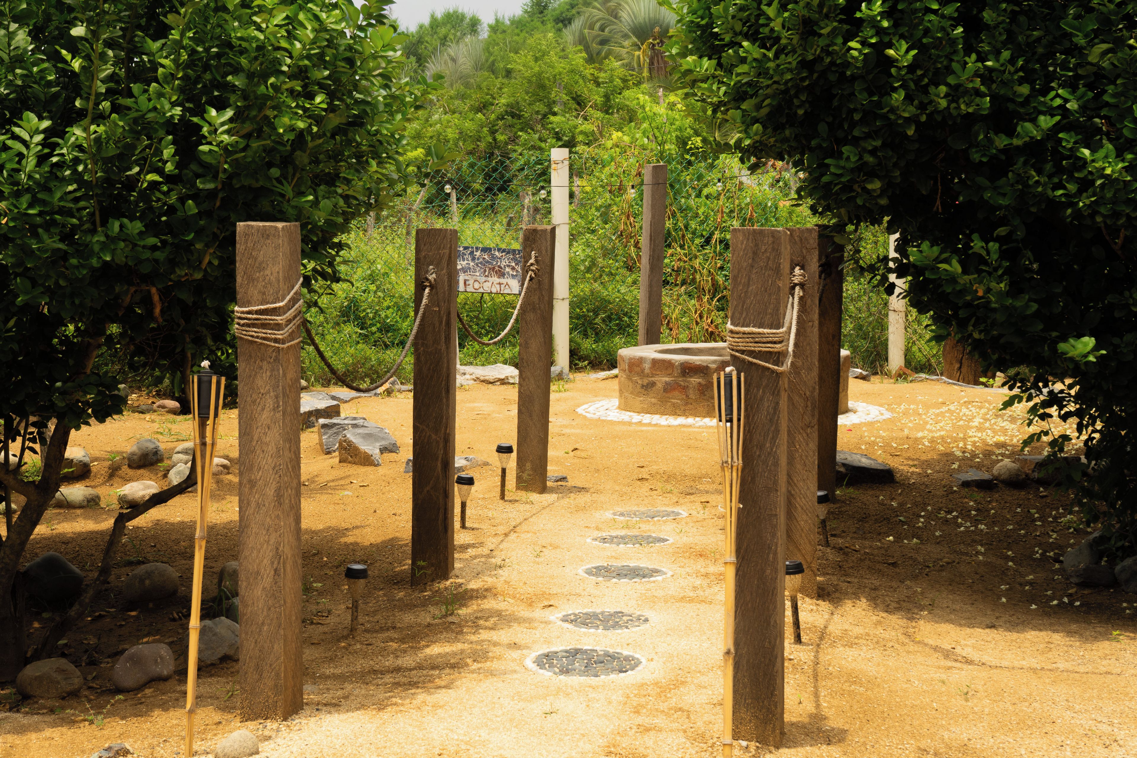 Un sentier bordé de poteaux en bois et de cordes, qui mène à une structure circulaire en pierre, entourée de verdure.