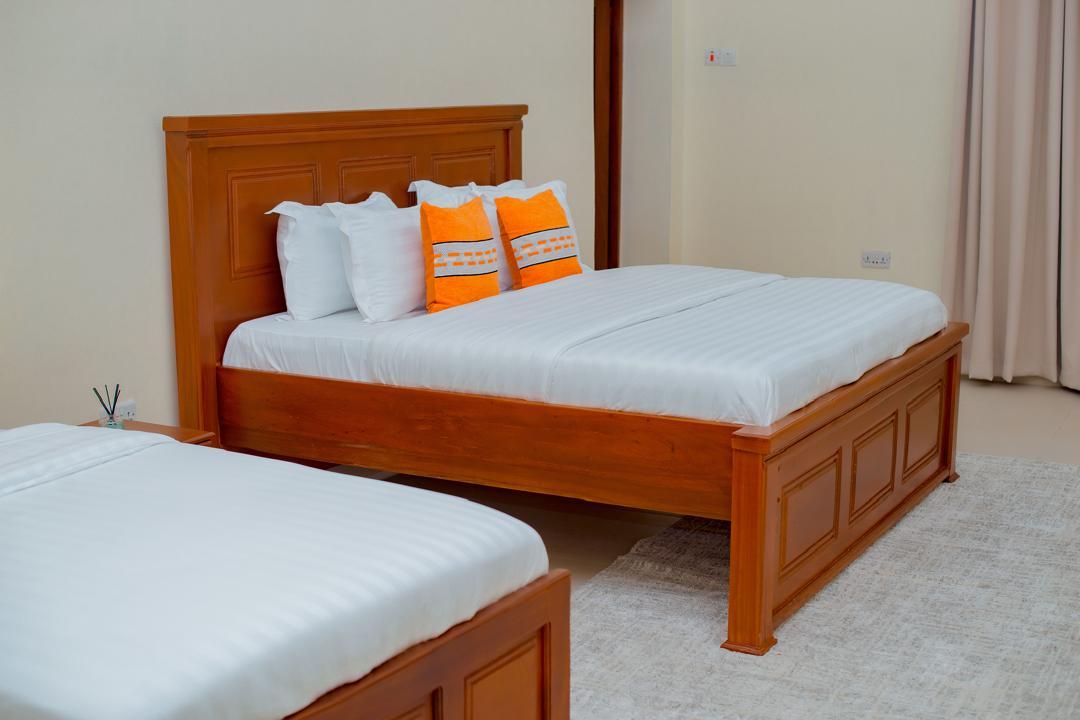 A double bed with a wooden headboard and two pillows, one white and one orange, in a room with a beige carpet.