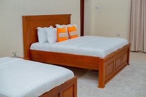 Un lit double avec une tête de lit en bois et deux oreillers, un blanc et un orange, dans une pièce dont le sol est recouvert d’une moquette beige.