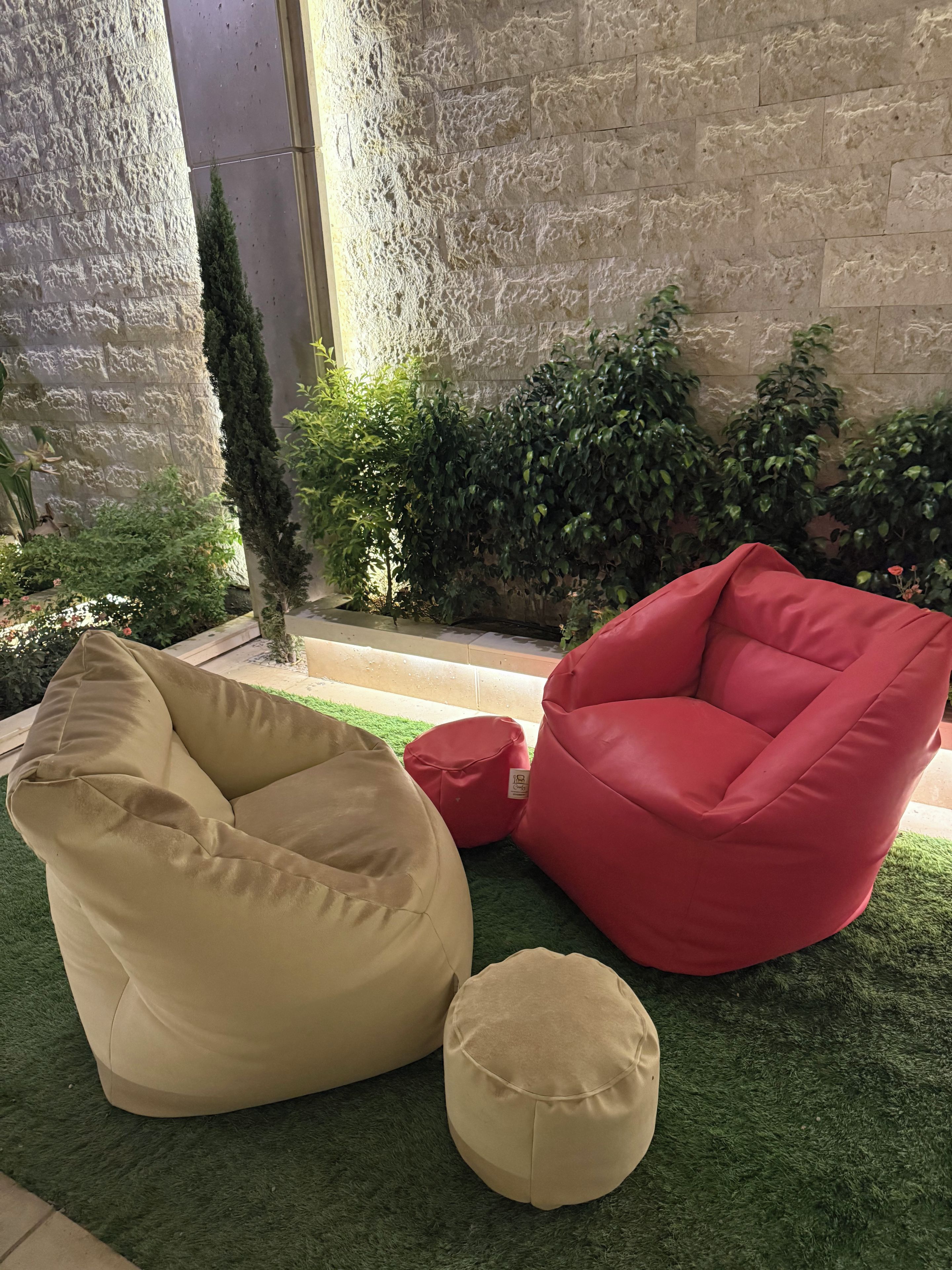 A beige bean bag chair, a red bean bag chair, and a small round ottoman on artificial grass.