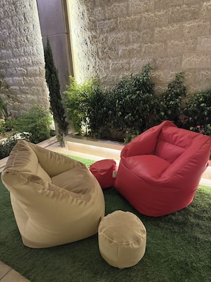 Un fauteuil en forme de sac de haricots de couleur beige, un autre de couleur rouge, et un petit pouf rond posé sur de l’herbe artificielle.