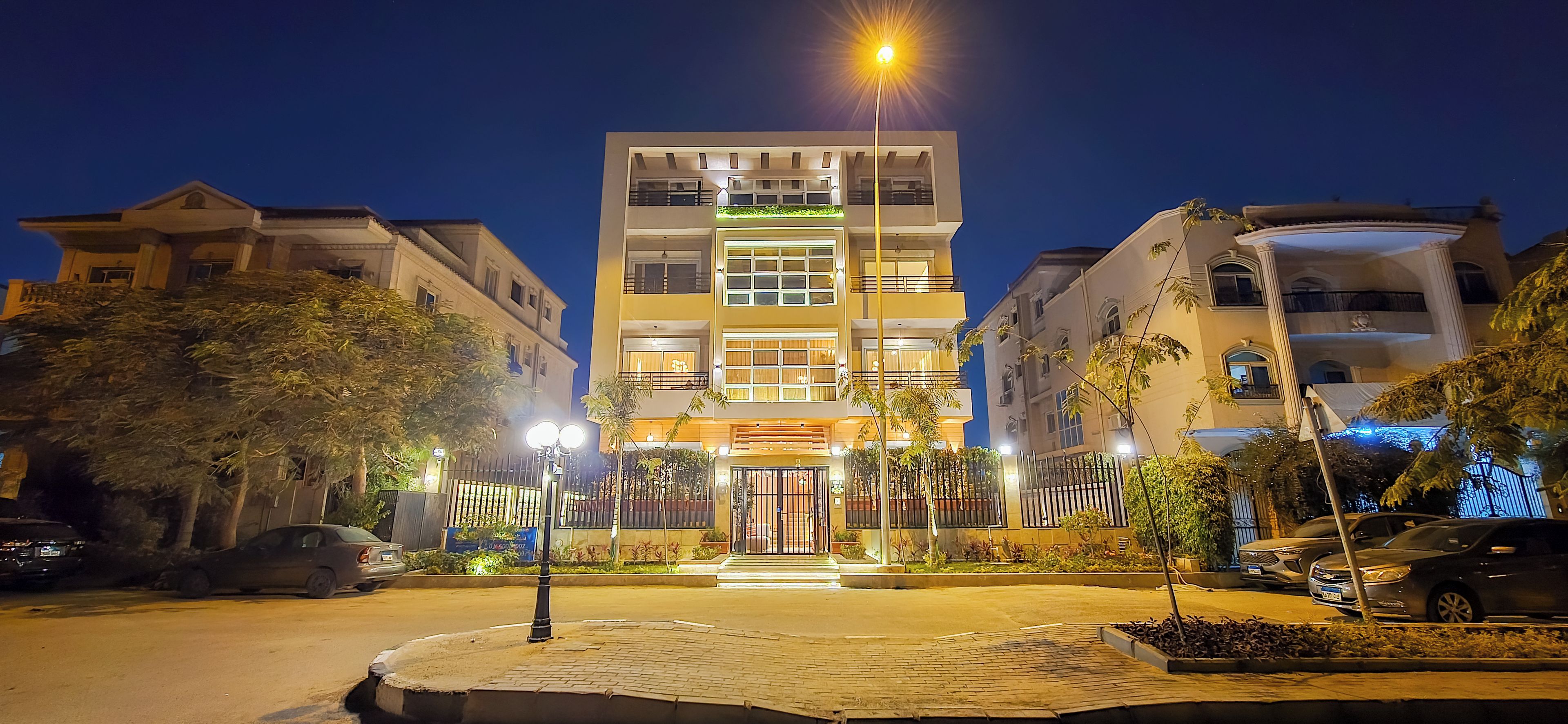 A multi-story hotel with a gated entrance, illuminated by street lamps, surrounded by parked cars and trees.