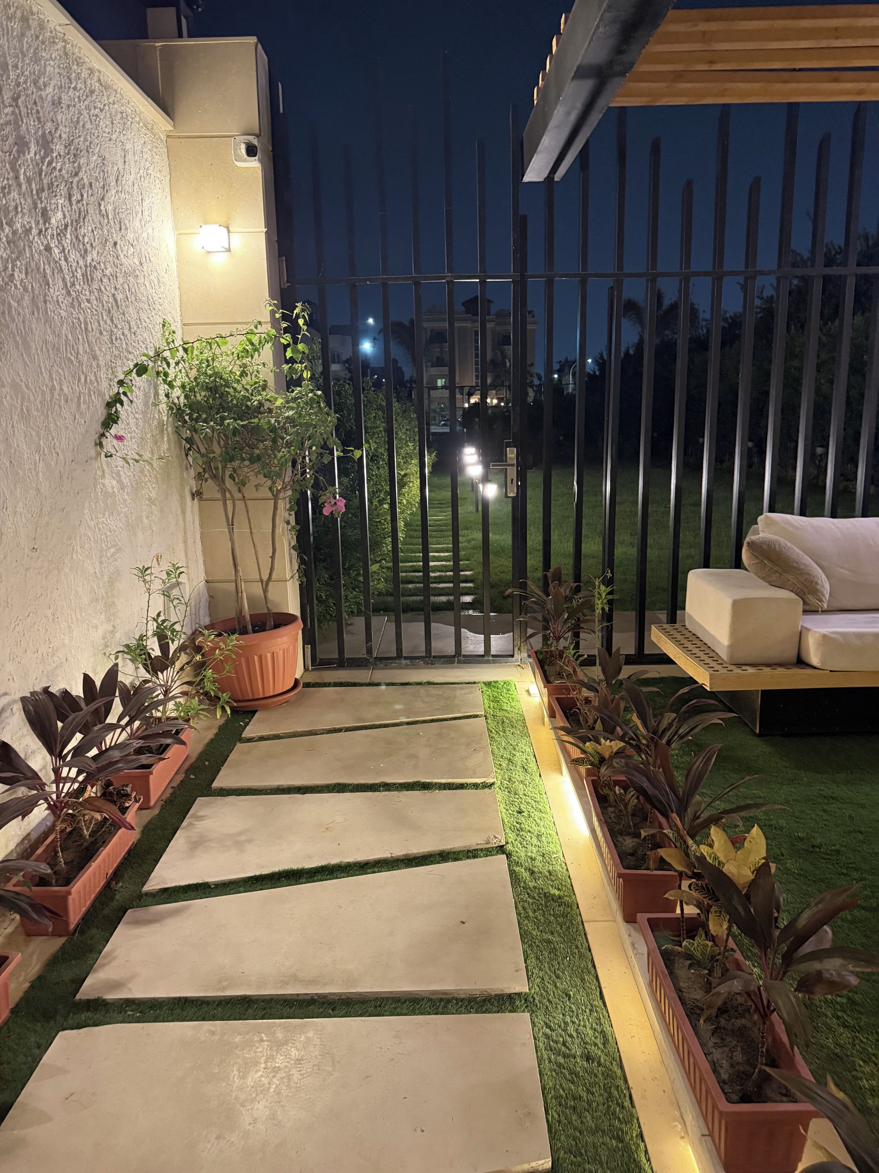 A paved pathway leading to a gated entrance, flanked by potted plants and a seating area with cushions.