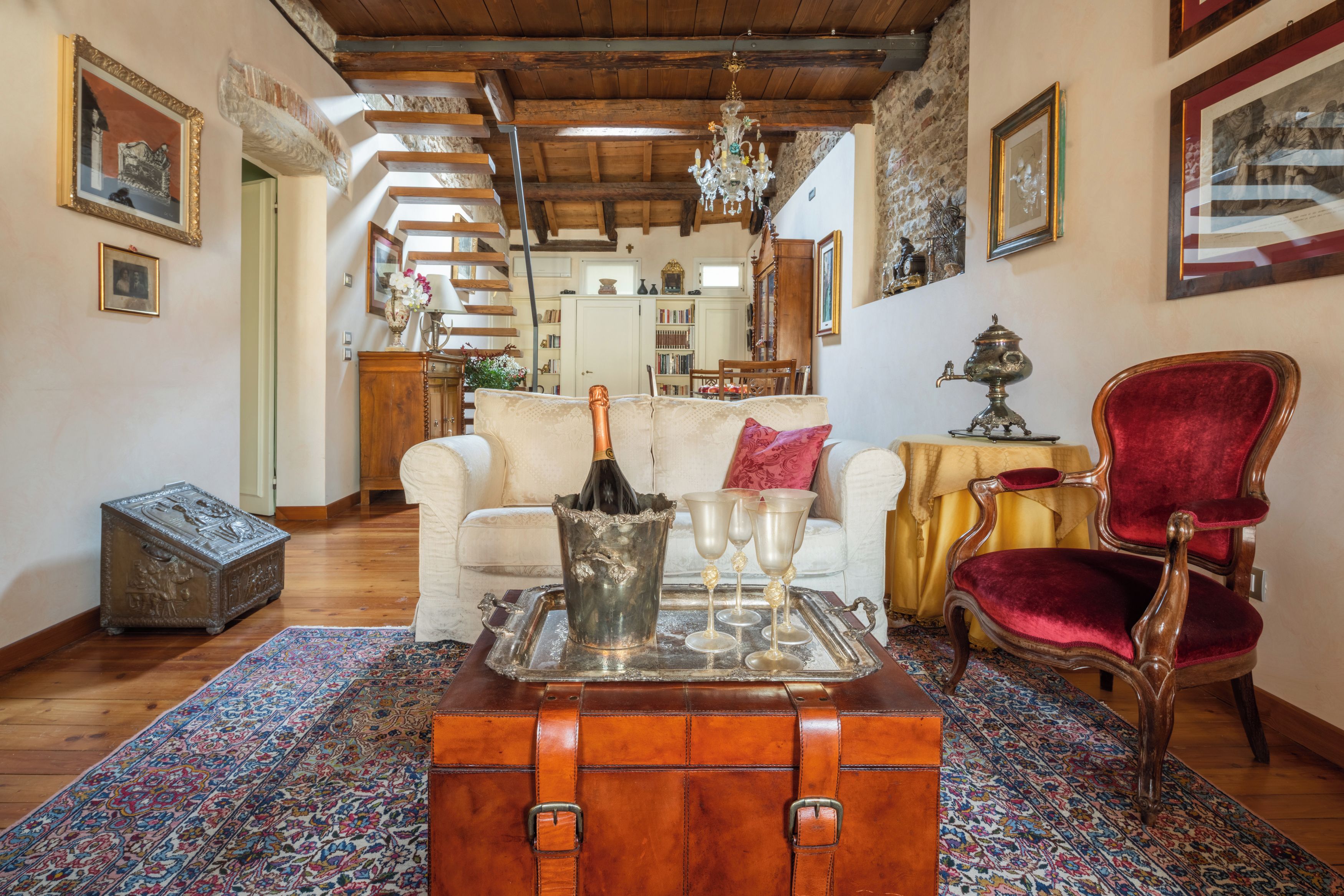 Un vaste salon doté d’un plafond en bois, d’un tapis à motifs, d’une malle ancienne, d’un fauteuil en velours rouge et d’un lustre.