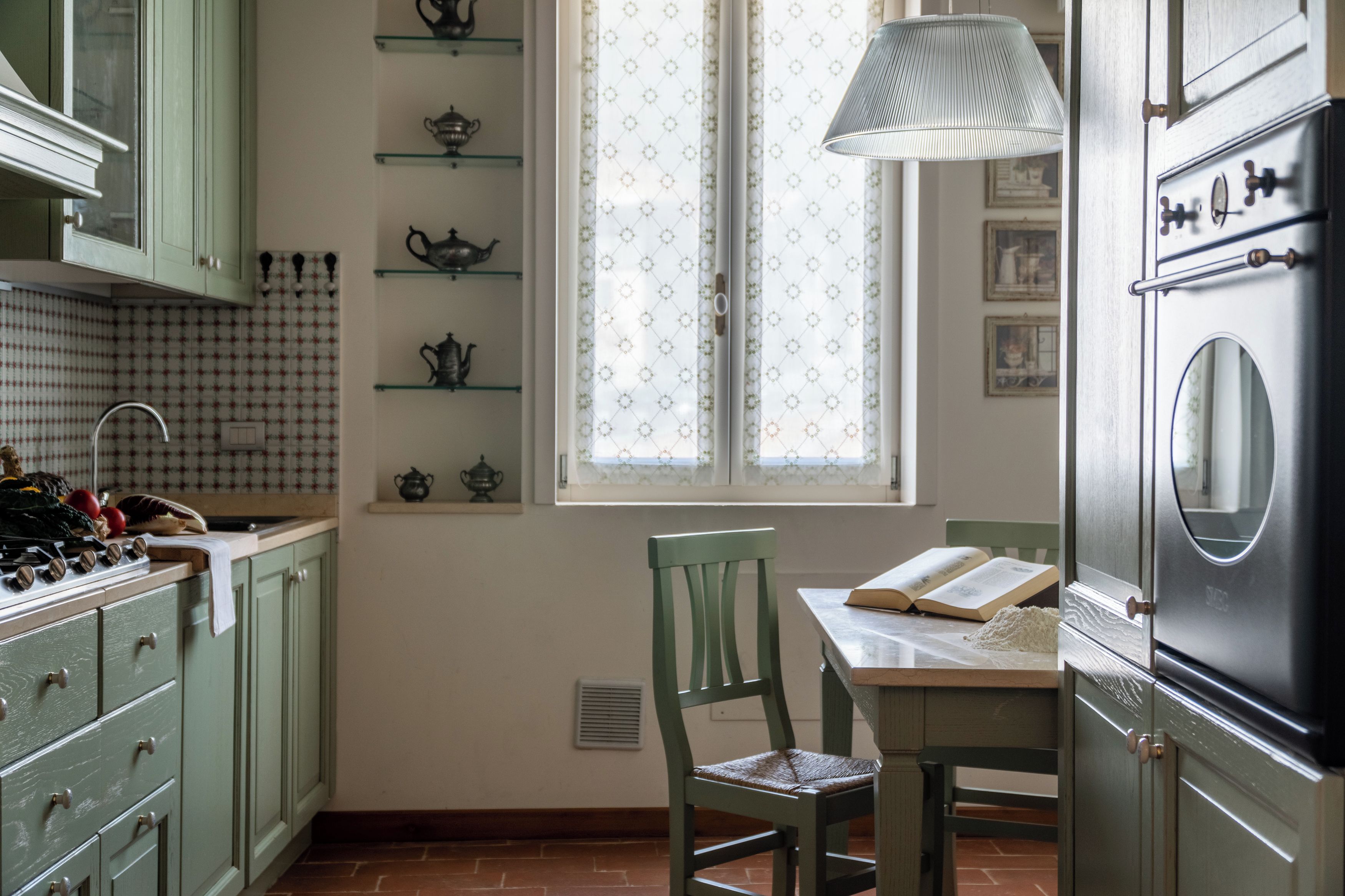 Una cucina con mobili verdi, un fornello, una finestra con tende e un tavolo con una sedia.