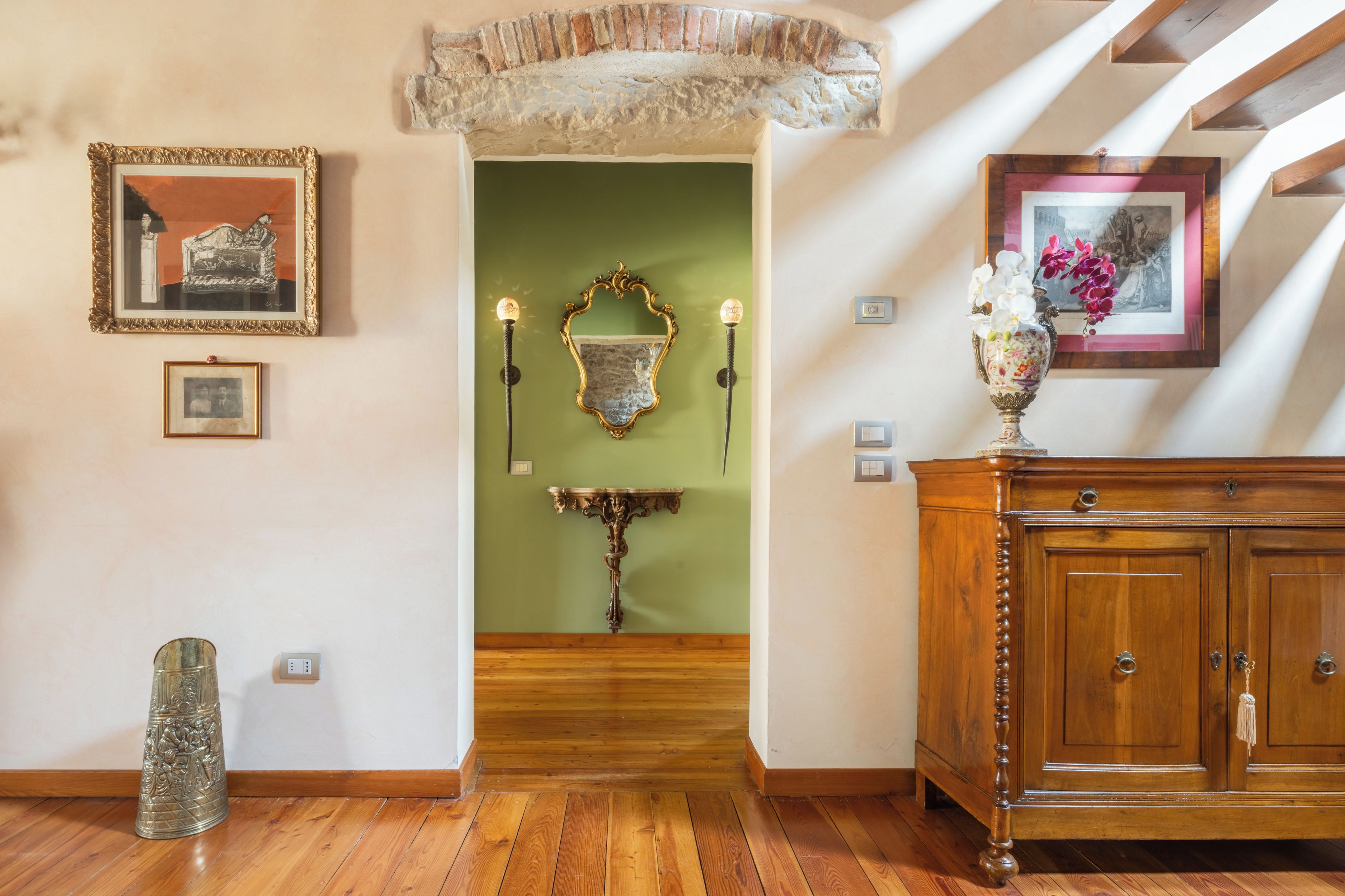 A hallway with a green wall, wooden floor, a mirror, a decorative wall piece, and a cabinet with a vase.