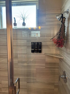 A shower area with a glass door, wooden wall tiles, a hanging showerhead, and three bottles of body wash. - Spa-Inspired Escape Cozy 2BR/2BA with Private Yard in Fort Worth (Fort Worth)