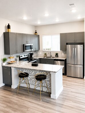 A modern kitchen with grey cabinets, stainless steel appliances, a marble countertop, and two bar stools. - Spa-Inspired Escape Cozy 2BR/2BA with Private Yard in Fort Worth (Fort Worth)