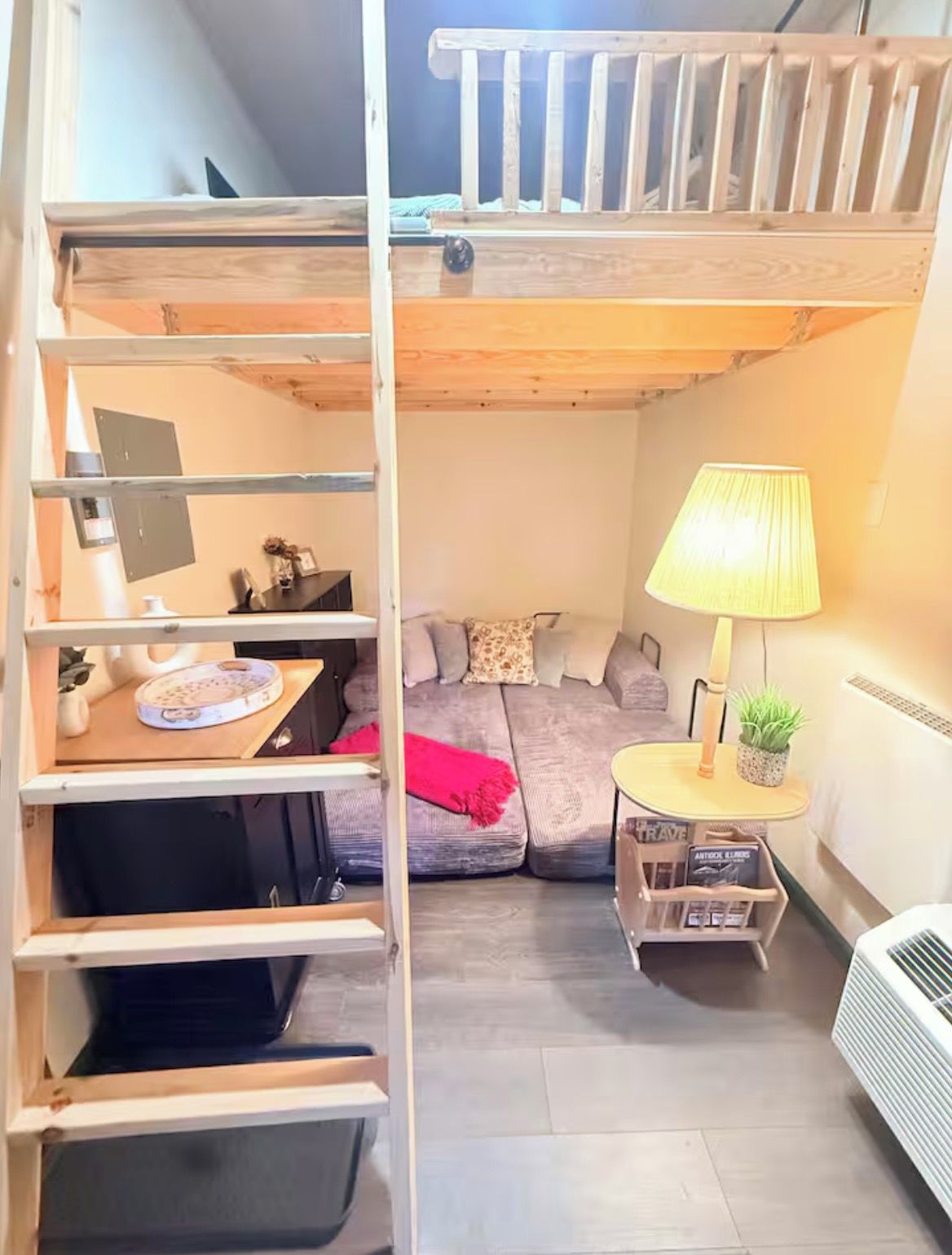 Een kleine kamer met een stapelbed, een bureau met lamp, een tafeltje met een plant en een bank.