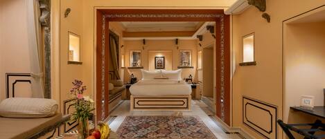 Quarto de hotel com uma cama grande, uma mesinha com frutas, um espelho decorativo e vista para o corredor.