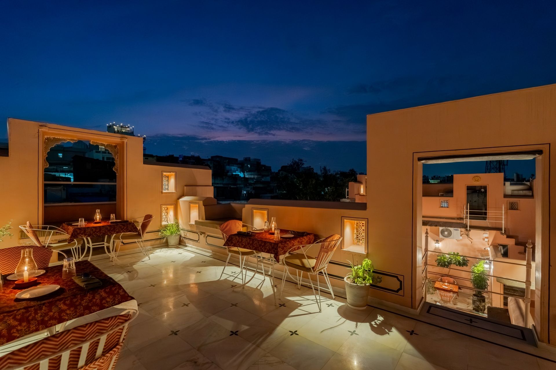 Una terraza en la azotea con mesas y sillas, plantas en macetas y vistas panorámicas de la ciudad al atardecer.