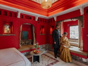 A room with red walls, a bed, a window with curtains, and a small table with fruit. - Indulge Udaipur (Phool Haveli) by PACK (Udaipur)