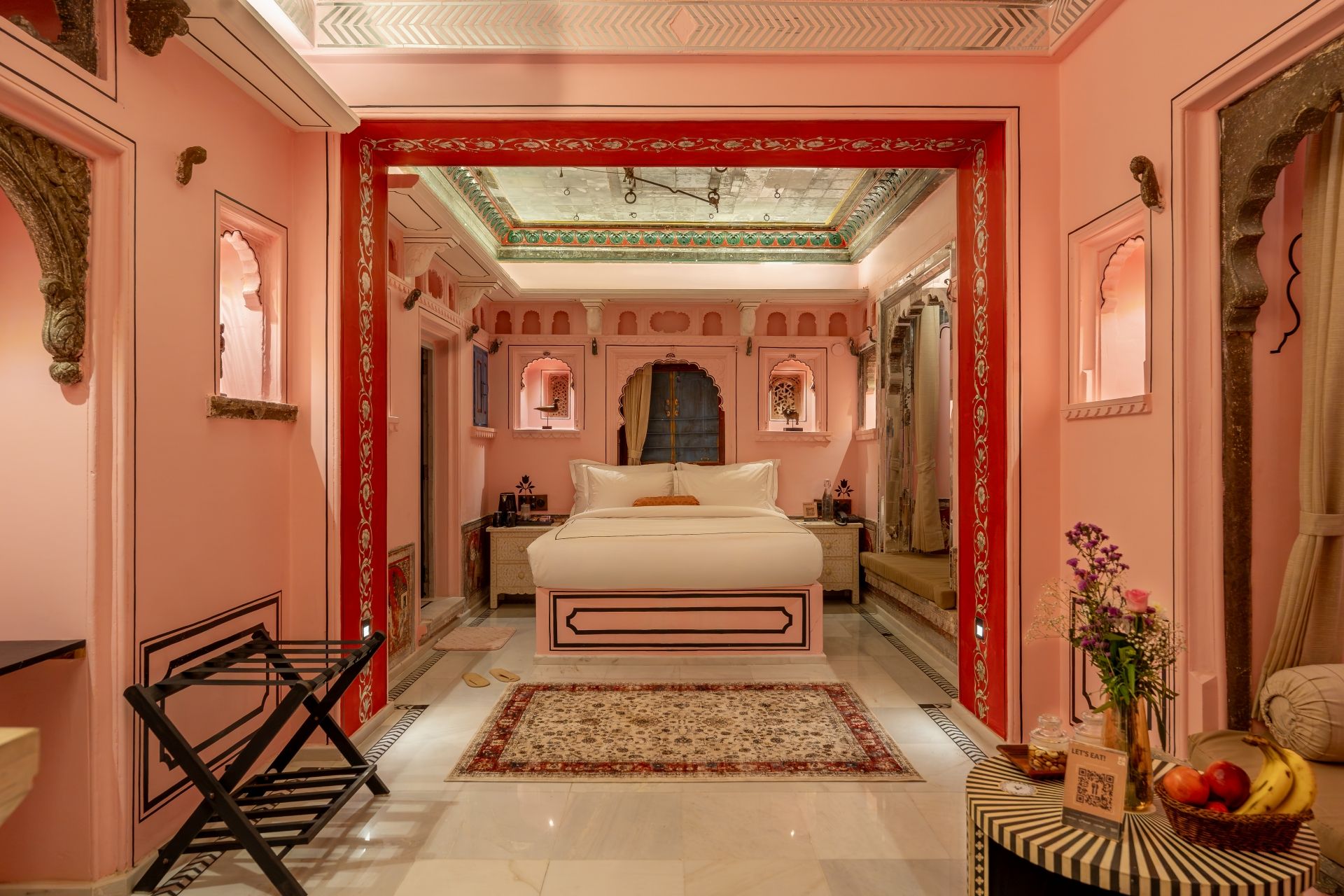 Habitación de hotel en tonos rosados, con cama, mesita de noche, un arco decorativo rojo y vistas al pasillo.