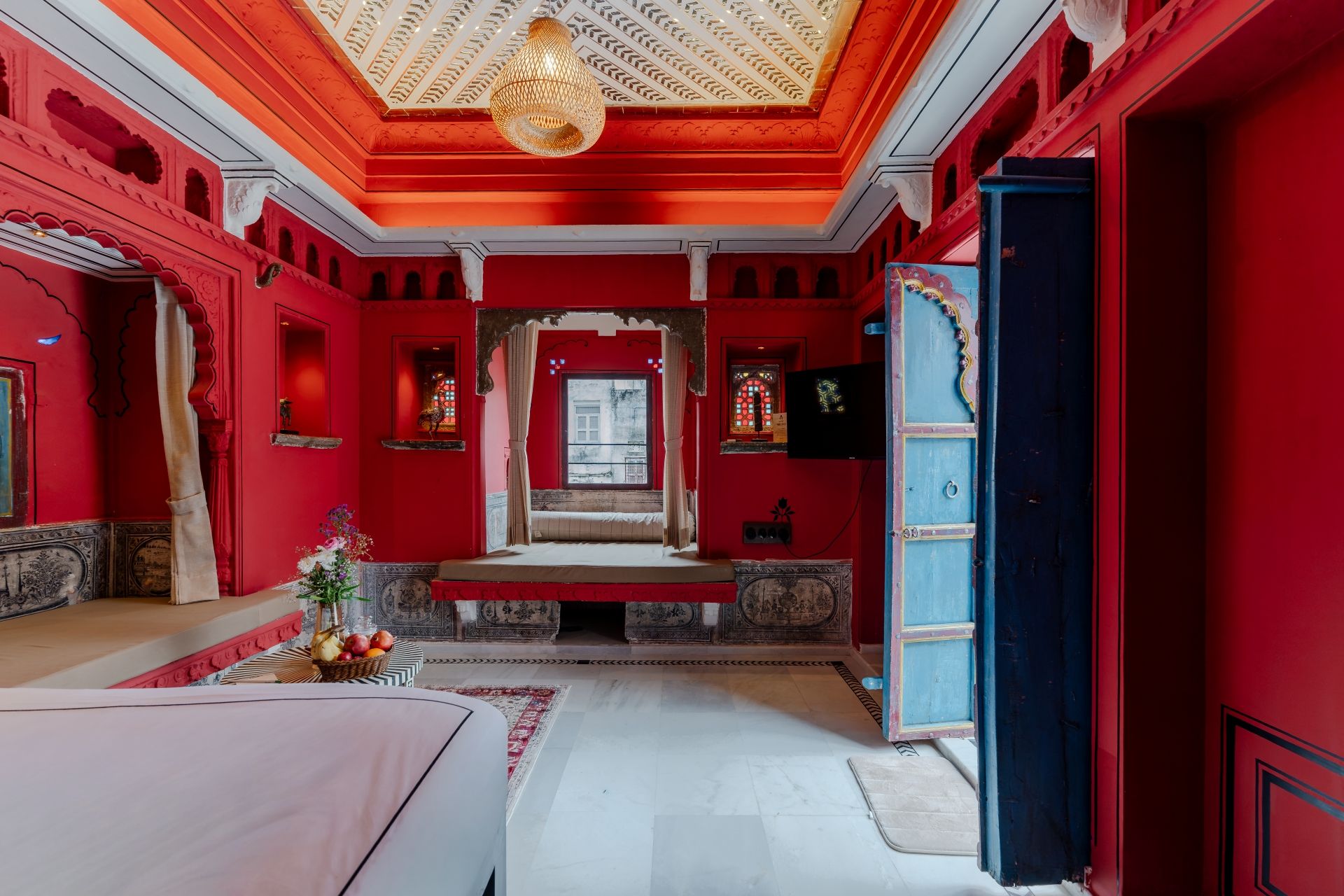 A room with red walls, a large bed, a chandelier, and a view of the street.