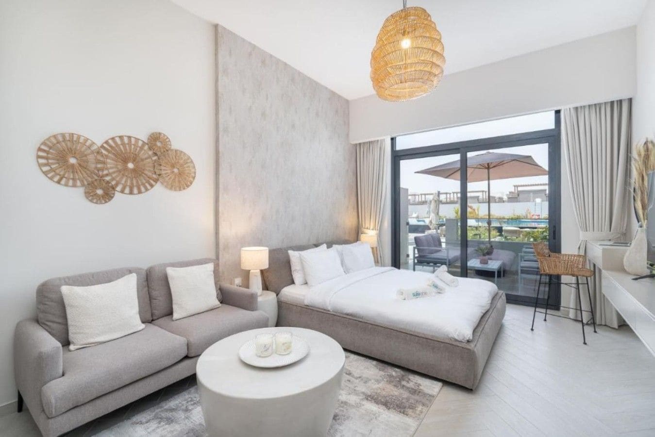 A modern hotel room with a large bed, a sofa, a round coffee table, and a balcony with outdoor seating.
