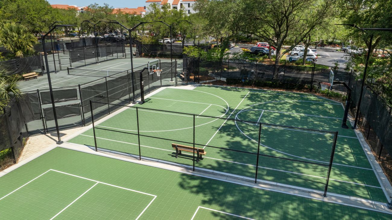 An outdoor basketball court with a wooden bench, surrounded by a black fence and trees.